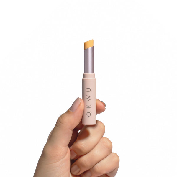 Mano sosteniendo el labial OKWU Magic Lipstick abierto, con su barra amarilla pH-reactiva y envase rosa